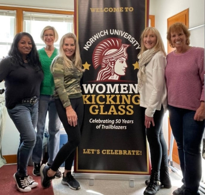 Five women standing indoors in front of a large Norwich University Women Kicking Glass banner, smiling and posing together.